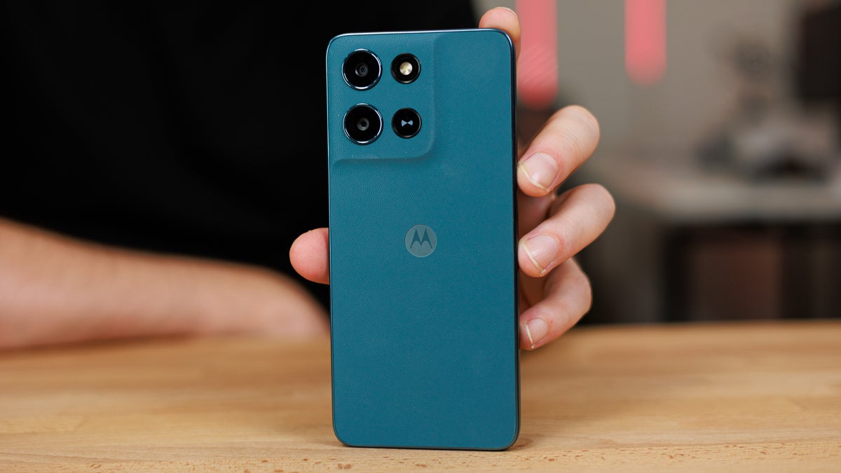 Hand holding green Motorola phone upright on a wooden table, back panel facing camera..